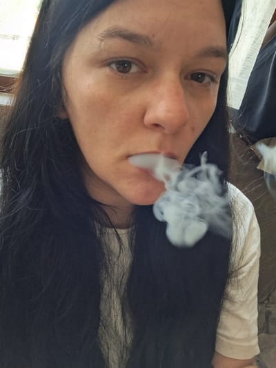A close-up, indoor shot shows a person exhaling smoke. The person has dark hair and is wearing a light-colored shirt. Smoke is visible around their mouth and nose, dissipating into the air. The background is blurred but suggests an interior setting. There is no visible indication of the location being in Marchevo, Bulgaria.