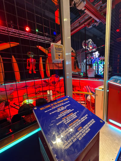 The image depicts the interior of an arcade or amusement venue, likely in Panama City Beach, United States of America, based on the context of similar venues in that location. The focal point is a large, blue sign for "THE HUMAN CRANE" game, which is partially visible and displays safety rules and operational instructions. The game itself appears to be a claw or crane machine designed to pick up prizes.
In the background, through a black metal grate, various arcade games and attractions are visible, illuminated by vibrant red and blue lights. Some of these include digital scoreboards showing numbers like "50" and "25.0". Several people are present within the venue, though their faces are not clearly distinguishable due to the lighting and distance. One individual in a white shirt is operating one of the games. In the foreground, a stuffed alien figure in red clothing is suspended, presumably as a prize or part of the game's display. The overall environment is characterized by bright, artificial lighting and the typical clutter of an entertainment arcade.