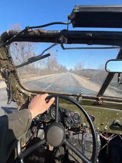 A first-person perspective photograph captured from the driver's seat of a heavily used, olive-drab vehicle traveling on an asphalt road in Voronino, Russia, under a clear blue sky. A fair-skinned human hand, wearing an olive-drab sleeve, is visible gripping the top of a black, three-spoke steering wheel.
The vehicle's interior components visible include a worn olive-drab metal dashboard with several round gauges (speedometer, tachometer, fuel/temperature), some switches, and bundles of exposed electrical wiring. Keys with a blue lanyard and a round keychain are in the ignition. The windshield is extensively splattered with dried mud and dirt, partially obscuring the view, with two windshield wiper blades positioned upwards. The windshield frame is olive-drab metal, exhibiting significant rust and damage, particularly on the driver's side. A black rectangular LED light bar is mounted externally above the windshield. A small, black-framed rearview mirror with a dangling red, yellow, green, and black beaded decoration is visible on the right side of the windshield. A silver-colored wrench rests on the dashboard near the passenger side windshield base.
The exterior scene depicts a two-lane asphalt road extending into the distance, bordered by dirt shoulders and sparse forest lines of bare deciduous trees on both sides. A power line tower is visible in the distant background on the right. Distant vehicles are faintly discernible on the road ahead. The clear sky suggests fair weather.