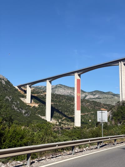 The image captures a vast concrete viaduct spanning a mountainous, vegetated valley in Gornje Mrke, Montenegro, under a clear blue sky. The multi-pier bridge, light gray in color, extends from the left mid-ground towards the right, supported by numerous tall, rectangular pillars. A prominent feature is a large, vertical red banner displaying the golden Montenegrin coat of arms affixed to one of the central bridge piers, extending nearly its full height. The surrounding landscape consists of steep, rocky hillsides densely covered in green shrubs and trees, with more distant mountains visible in the background. Shadows from the bridge are cast across the distant terrain. In the immediate foreground, a paved road with a metal guardrail featuring white reflectors runs along the lower edge of the frame. Behind the guardrail on the right, a gray square sign with a white house symbol and an upward arrow is mounted on a pole. Two small, dark specks, one resembling a bird, are visible in the bright blue sky. No people are present.