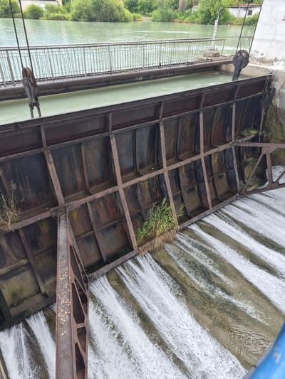 This image shows a dam structure on a river. The dam is made of rusted metal panels and supports, with water flowing over it in multiple streams, creating a cascading effect. A metal walkway with a railing is visible above the dam. In the background, there is a body of water, lush green vegetation, and a building. The dam appears to be part of an infrastructure in an outdoor, possibly rural or semi-urban, setting. The specific location in the city of Tekeli, Kazakhstan is not identifiable from the visual information provided in the image.