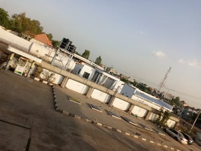 The image captures a street view in Hotoro North, Nigeria, with a focus on a fuel station and its surroundings. The perspective is slightly angled, looking down and across the scene. In the foreground, a concrete forecourt is visible, with patterned paving stones forming a walkway and some recessed rectangular covers. Beyond the forecourt, a white building with a blue-roofed overhang serves as the main structure of the fuel station. Several fuel pumps with prominent branding are positioned in front of this building. Adjacent to the building are large, white cylindrical storage tanks, some of which are elevated and topped with black water tanks. A white fence with barbed wire runs along the perimeter of the fuel station's property. In the mid-ground, trees and other vegetation are present. Beyond the fuel station, a street with other buildings and a telecommunications tower can be seen. Two cars, a silver sedan and a darker colored sedan, are parked on the right side of the image. The sky is clear with a pale blue hue, suggesting it is daytime. A few people are visible in the distance, walking near the street. The overall impression is that of a functional commercial area within a residential or mixed-use part of the city.