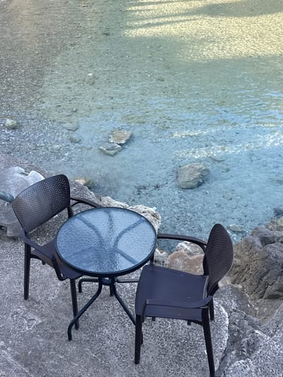 The image depicts a serene outdoor setting by a body of water in Seoca, Montenegro. In the immediate foreground, a rough, grey ground surface, consistent with concrete or paved stone, anchors the scene. Positioned on this surface are two dark-colored, possibly black or dark brown, outdoor chairs with perforated backs and a small, round table with a dark metal frame and a textured, translucent glass top. One chair is situated to the left of the table, angled towards it, while the second chair is on the right, also angled towards the table. No individuals are present at or near the table.
Beyond the seating arrangement, the terrain transitions into a rocky shoreline composed of varying sizes of grey and brown stones. This leads into a body of exceptionally clear, shallow water. The water's surface is lightly rippled, reflecting sunlight in various patterns, and its transparency allows for an unobstructed view of the underwater environment. The bottom is covered with numerous small, light-colored pebbles and several larger, irregular rocks. Distinct shadows are cast across the upper section of the water, suggesting overhead structures or foliage not visible within the frame. The overall impression is one of tranquility and natural beauty.