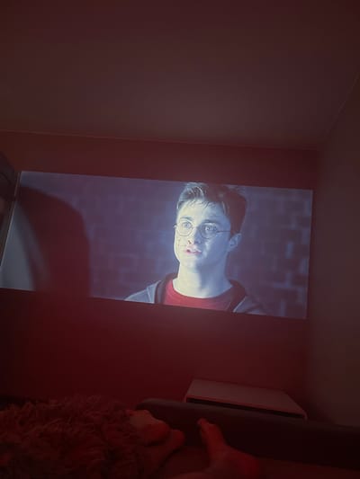 The media file is a photograph taken in Gliwice, Poland. The primary subject is a projection of a scene from a Harry Potter movie, featuring the character Harry Potter himself. He is shown from the chest up, with a close-up on his face. Harry is wearing his iconic round glasses and has a somewhat distressed expression, with a bloody nose. The background of the projection is a dark, brick-like texture.
The projection is being displayed on a wall in a dimly lit room. To the right of the projection, a white projector or media device is visible. Further to the right and slightly out of focus, a person's foot and a furry blanket or rug are present. The room itself appears to be a domestic setting, likely a living room or bedroom, given the presence of a projector and soft furnishings. The lighting is predominantly red, suggesting ambient light from the room or possibly a light source within the camera's vicinity. The image is oriented in portrait mode, with the projection appearing upside down relative to the typical viewing orientation. No text is visible in the media file. The overall mood conveyed is one of relaxed, at-home entertainment, with the intensity of the movie scene contrasting with the casual setting.