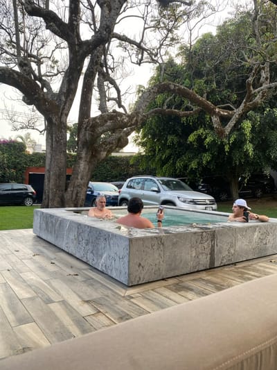 Three people are in a pool. A person wearing a white cap holds a phone in the pool water. Another person has their back to the camera. An older man is visible in the pool. Cars are parked beside the pool. A large tree with many branches is beside the pool. A portion of a chair is visible in the bottom right corner.