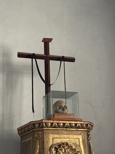This image depicts a wooden cross with two black ribbons hanging from it, placed atop an ornate, gold-colored pedestal. In front of the cross, a human skull is displayed inside a glass case, resting on a wooden block. The background is a textured, light-colored wall. The scene is located in the San Miguel Arcángel church in Huejotzingo, Mexico, likely within a reliquary or devotional display.