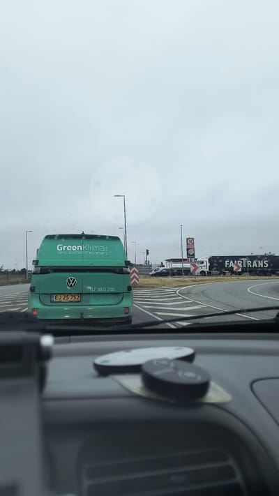 The image was taken from inside a vehicle on a cloudy day in Ølby, Denmark. The vehicle is stopped at an intersection, with a green Volkswagen van in front. The van has a license plate that reads "EJ 25 752" and the words "Green Klima" and "Ventilationsentreprenør" are visible on its rear door, along with a phone number "TLF: 8173 0199". To the right of the van, there is a circular intersection with a large tanker truck that says "FAIRTRANS". In the background, a Burger King and a Circle K gas station are visible. The dashboard of the vehicle in the foreground shows two round objects, possibly car remotes or key fobs.