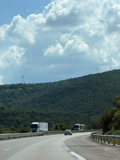 The image depicts a roadside scene with a highway winding alongside a densely forested mountainside. Several vehicles are visible on the road, including a white truck with a silver trailer, another white truck, and a white car. The trucks appear to be in motion, suggesting a travel or transport activity. The mountainside is covered in lush green trees, indicating a natural and possibly rural setting. The sky is partly cloudy with dramatic white clouds set against a blue sky. Electrical pylons and wires are visible on the mountainside, hinting at infrastructure present in the area. The road has a white line marking the lanes and a metal guardrail separating it from the embankment. The presence of the road and guardrail suggests a developed area, likely a highway, facilitating travel through this mountainous region. The lighting suggests it is daytime, possibly late morning or afternoon, given the brightness of the sky and the shadows. There are no people explicitly visible in the image. One of the trucks has a small green sign with the number "23" on it. The location context provided is Churek, Bulgaria.