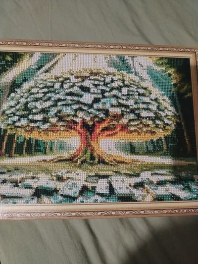 The image shows a framed diamond painting depicting a tree with a lush canopy of white and green, set against a backdrop of trees and foliage. The tree's trunk is a vibrant mix of red and orange. The foreground features a rocky, watery landscape. The painting is presented on a green, textured surface, possibly a tablecloth or fabric. The frame of the painting is ornate and gold-toned. There is no information in the image that indicates its location in Taldykorgan, Kazakhstan.