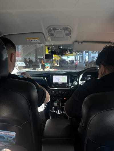 Inside a vehicle, two people are visible. The driver is seated in the front with their hands on the steering wheel, while the passenger is in the back. The vehicle features a navigation display screen on the dashboard. Outside the window, a blurred scene of people and buildings suggests a public or commercial area. The interior is predominantly black, with lighter shades on the ceiling and window frames.