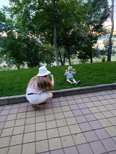 In Kelermesskaya, Russia, on a paved walkway and grassy incline, a person wearing a white bucket hat and a light pink shirt with beige shorts is crouched down, holding a phone to take a photo. The person's back is to the camera, and their long, brown hair is visible. In front of them, a young boy in a gray t-shirt and green shorts sits on the grass, wearing a light green cap and holding a stick in front of his face. The background consists of lush green trees and a body of water in the distance. The ground in the foreground is paved with square, interlocking tiles in varying shades of beige and grey, with shadows cast across them.