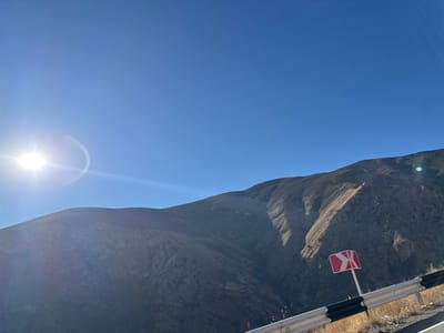 A bright sun shines in a clear blue sky, illuminating a large, barren mountain. The mountain's slopes are carved with deep crevices, and the terrain appears dry. A road sign with red and white chevrons stands next to a guardrail in the lower right corner, suggesting a winding road. The landscape evokes a sense of remoteness and rugged beauty.