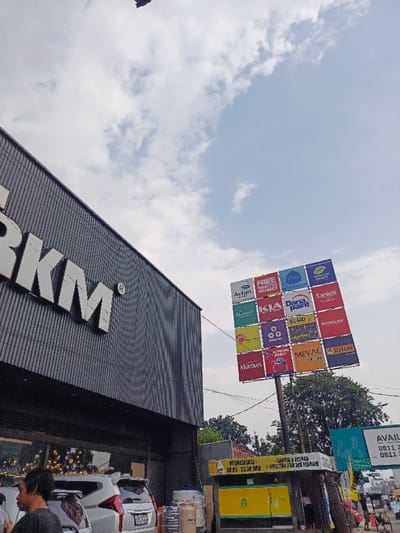 A multi-branded advertising signpost showcases various paint brands like Avian, Dana Paint, Sanlex, Nippon Paint, and Jotun. The sign is positioned near a building with "RKM" on its facade. A yellow and green booth displaying operational hours is visible at the signpost's base. Cars are parked in front of the RKM building, and trees are in the background under a partly cloudy sky. A black object is visible in the top of the image.
