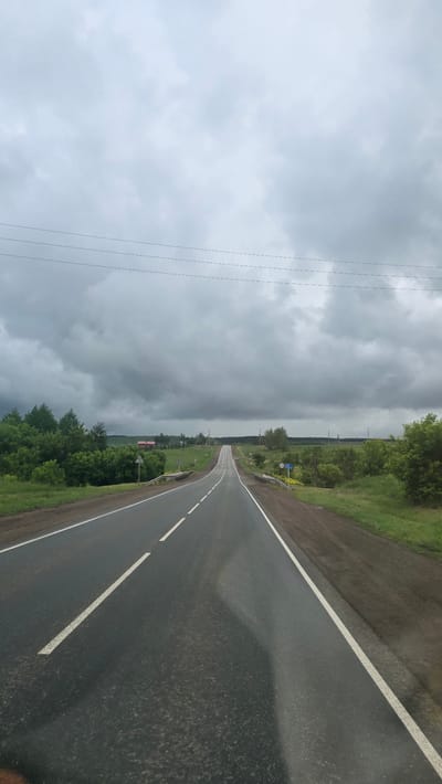 A two-lane asphalt road with a dashed white center line and solid white shoulder lines stretches into the distance, appearing wet under a heavily overcast sky dominated by dark grey clouds. Utility lines with multiple strands traverse the upper portion of the frame, running parallel to the road. On the left side, dense green trees and bushes border the road, with a red-roofed building partially visible beyond them. A white guardrail lines the roadside where it begins a gentle descent. On the right, green vegetation, including scattered trees and fields, extends outwards, also featuring a white guardrail and a blue informational road sign. The road itself ascends over a crest in the far background, framed by rolling green terrain under the cloudy horizon. No people or vehicles are present in the scene, which depicts a rural landscape near Кама-Исмагилово, Russia.