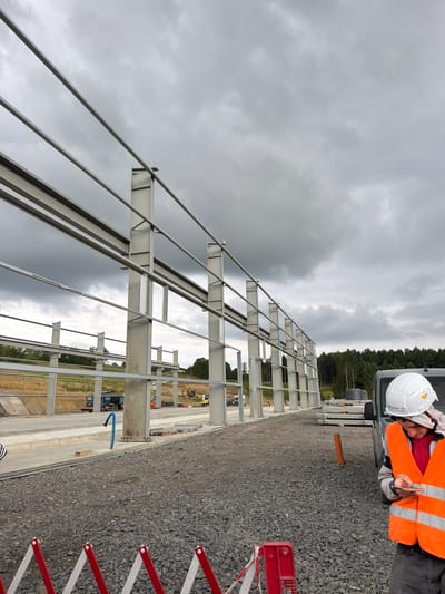 A man wearing a white hard hat and an orange safety vest stands on a gravel surface, looking at his phone. He is partially obscured by a dark gray vehicle to his right. Behind him, a large, unfinished steel structure with visible beams and columns dominates the frame. The sky is overcast with dark clouds. In the background, there is a wooded area with a line of trees and a construction site with heavy machinery, including trucks and excavators. The image appears to have been taken outdoors in Stróża, Poland, as indicated by the context.