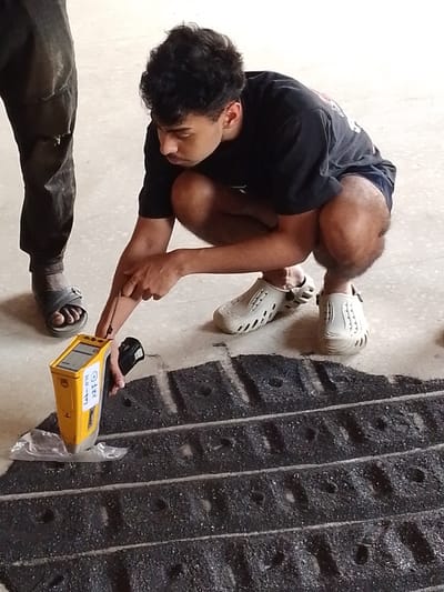 In Oko Erin, Nigeria, an indoor scene on a light-colored concrete floor depicts a young male subject, dressed in a dark t-shirt, dark shorts, and light slip-on shoes, crouched over a scientific setup. He is operating a yellow handheld X-ray fluorescence (XRF) analyzer labeled "Lab-011 XRF" which rests on a clear plastic sheet. His right index finger points towards the device or the sample beneath it. Directly below the analyzer and plastic sheet is a dark, granular material arranged in several parallel rows, each containing small, circular indentations. The lower leg and foot, clad in dark sandals and torn dark trousers, of a second individual are visible on the left side of the frame, standing adjacent to the primary subject.