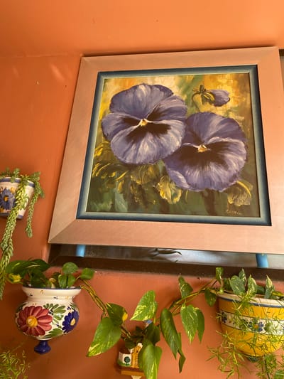 A framed painting of two purple pansies is displayed on an orange wall. The painting, which appears to be oil on canvas, has a light pink frame and a blue inner matting. A signature, "Schöttler," is visible in the upper right corner of the painting. Hanging plants are present on either side of the painting. To the right, a vibrant yellow ceramic pot, decorated with blue flowers, holds a trailing plant. Below this, another ceramic planter, adorned with painted flowers in red, blue, and yellow, also supports a trailing plant. To the left of the painting, a thin, light-colored vertical element is visible. The overall impression is of an outdoor or semi-outdoor space, possibly a patio or balcony, decorated with art and plants. The lighting suggests daytime.
