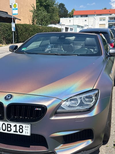 The image shows a close-up of the front of a chameleon-colored BMW M6 convertible parked on a street in Biberach an der Riß, Germany. The car's hood and front bumper exhibit a rainbow-like iridescent effect. The vehicle has a black grille with an "M6" badge on the right side, and a "G-POWER" emblem on the left. A German license plate, "OB 18 03/11", is visible below the grille. In the background, a bus stop sign with a yellow "H" symbol and informational panels is present, along with a neatly trimmed hedge, buildings with red and white facades, and other parked vehicles. The sky is clear and blue, indicating a sunny day. No people are visible in the image.