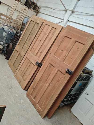 The image depicts three wooden doors leaning against a wall in an indoor storage space. The doors are of varying shades of brown, with a panelled design and black hardware. In the background, there are chairs, a metal cage, and other miscellaneous items. The walls appear to be white, possibly brick or concrete. The location appears to be a warehouse or storage unit in Swindon, United Kingdom. There are no people visible in the image.