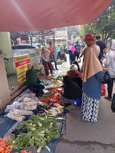 A bustling outdoor market scene displays a vibrant array of vegetables on a blue tarp, including carrots, bok choy, eggplants, and chilies. Several people gather around the spread, some squatting to select their produce while others stand and observe. A red awning provides shade from the sun. Nearby is a sign advertising rentals. Motorbikes and pedestrians fill the background, creating a lively urban setting.