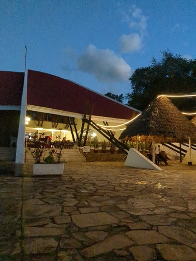 The image shows an outdoor patio area of a restaurant at dusk in Morogoro, Tanzania, located at coordinates -6.835 latitude and 37.672 longitude. The patio has a stone floor and is surrounded by a large red-roofed building on the left and a thatched roof structure on the right. There are a few trees and bushes scattered around the patio. String lights are strung up around the patio, providing illumination. The thatched roof structure appears to have a small entrance with a person sitting on a chair in front of it. The red-roofed building has a large open space with several tables and chairs set up for dining. There are people inside the restaurant sitting at the tables. The sky is blue with a few clouds. The image was likely taken at night with the artificial lights from the patio and the restaurant providing illumination.
