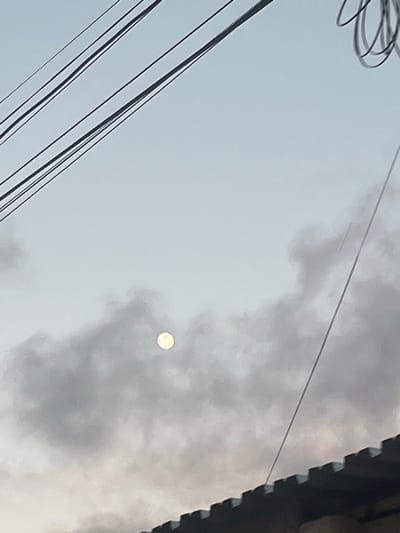 The image shows a hazy sky with the moon visible as a pale circle, partially obscured by clouds. Several power lines crisscross the upper portion of the frame. In the foreground, the edge of a dark, tiled roof with a jagged silhouette is visible. The scene is likely set in an outdoor urban environment, potentially in San Antonio, Venezuela, given the presence of power lines and residential roofing typical of the area. There are no people or specific actions depicted. The overall atmosphere is muted and somewhat overcast.