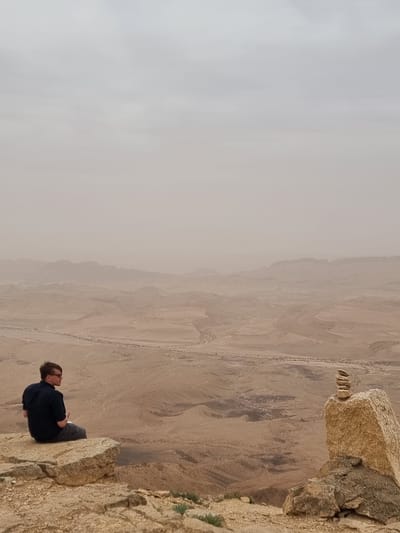 A man is sitting on the edge of a cliff overlooking a desert landscape. The sky is overcast. The location is Mitzpe Ramon, Israel. The man is wearing a dark shirt and dark pants. He is sitting with his legs crossed and his arms folded. The cliff is made of light-colored rock. There are some small plants growing on the cliff. The desert landscape is brown and tan. There is a road visible in the distance.
