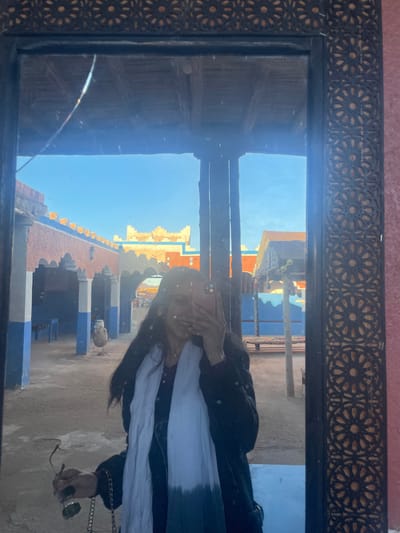 A mirror reflects a scene outdoors. The reflected image includes a person covering their face, wearing a white scarf and black jacket. They are holding sunglasses in their hand. The background shows a rooftop with blue and orange structures under a clear blue sky. The mirror itself has an ornate wooden frame with a geometric pattern. A crack is visible on the mirror's surface in the bottom left corner.