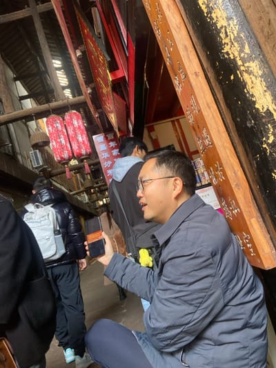 A man with glasses wearing a gray jacket looks at his phone. Several people surround him, all indoors. Red lanterns and wooden structures adorn the setting, suggesting a temple or traditional building. Carved wooden beams with Chinese characters run along the ceiling. The angle of the image is slightly tilted.
