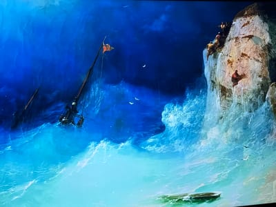 The image presents a dramatic coastal landscape, identified as being in the city of Reus, Spain. A turbulent sea, colored in gradients of deep blue to light turquoise, occupies the left and central foreground. A damaged sailing vessel, characterized by a dark, partially submerged structure and a mast bearing an orange flag, is visible amidst large, white-capped waves on the left. Several individuals appear to be in the water near the wreckage. Multiple white birds fly above the agitated water. On the right, a towering, jagged cliff, predominantly light brown and white, ascends from the sea. At the cliff's summit, a cluster of approximately four individuals is observed, seemingly observing the scene below. A single person, dressed in reddish attire, clings precariously to the lower face of the cliff. The environment suggests a perilous maritime incident and human struggle for survival against severe natural conditions.