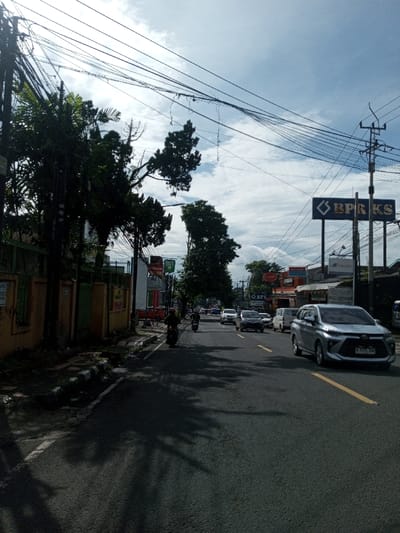 This image, captured in the city of Domphoog_Dungkarchhoeling, Bhutan, presents a daytime street scene with moderate traffic under a partly cloudy sky. On the left side of the asphalt road, a concrete sidewalk runs alongside a low, orange-brown wall, behind which dense green foliage and utility poles with a complex network of overhead wires are prominently visible. Two individuals, each riding a motorcycle and wearing dark clothing with a helmet, are traveling away from the viewer on the left side of the road.
On the right side of the road, a silver/light gray modern SUV, bearing an Indonesian license plate 'B 3072 TOG', is driving towards the viewer. Further along this side of the street, a large blue sign with white lettering 'BPR KS' and a diamond-shaped logo is displayed, supported by a pole. Adjacent buildings feature commercial signage, including 'BFI Finance' and 'APOTEK', indicating retail or service establishments. The asphalt road surface shows a solid yellow line near the right shoulder and a dashed white line delineating lanes further in the distance, accommodating a line of diverse vehicles moving in both directions. The sky above is characterized by patches of blue intermingled with white clouds, and shadows from unseen objects to the left are cast across the roadway.