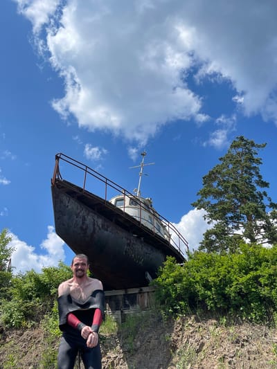 A man in a black and red wetsuit is lying on his side on a grassy, rocky embankment, smiling at the camera. He is positioned in the lower right quadrant of the image. Behind him, and taking up a significant portion of the frame, is a large, dark, rusted vessel, possibly a boat or barge, propped up on land. The vessel is angled upwards and to the left, with a rusty railing along its upper edge. Above the vessel, the sky is a clear blue with scattered white, fluffy clouds. Green trees and foliage are visible on the embankment and in the background, with a tall pine tree reaching towards the sky from the upper left. The scene appears to be outdoors on a sunny day. The man's expression is cheerful. There is no visible text in the image. The location context is Divnogorsk, Russia.