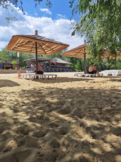 The image depicts a sandy beach scene under a partially cloudy blue sky. Two large, thatched umbrellas provide shade over several white lounge chairs. In the foreground, the sand is textured with shadows cast by foliage.

A man is seated on a lounge chair under the left umbrella, facing away from the camera. A toddler in an orange and white outfit sits on the sand to his left. Further back, a couple is seated on lounge chairs under the right umbrella, also facing away from the camera.

In the background, a long, low building with a thatched roof and a sign that reads "BOHO BEACH BAR" is visible. There are rows of empty lounge chairs in front of the bar. Trees and greenery surround the area, suggesting a natural or semi-natural setting.

The scene appears to be a recreational area, likely a beach club or resort, possibly located in Belaya Kalitva, Russia, given the context provided. The overall impression is one of leisure and relaxation on a sunny day.