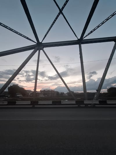 The image shows a view of a city on the horizon, seen through the latticework of a bridge, in Kungur, Russia. The sky is overcast with a sunset casting soft pink and orange hues. A multi-story building with a dark facade and many windows is visible in the center of the horizon, likely a prominent structure in the city. Trees line the banks of a river that flows through the cityscape. The bridge's metal framework is dark and industrial, framing the scene. The foreground consists of the asphalt road surface of the bridge, with a white line dividing lanes. A black and white striped barrier runs along the edge of the bridge. No people or vehicles are clearly visible. The overall impression is a tranquil, somewhat moody, urban landscape at dusk.