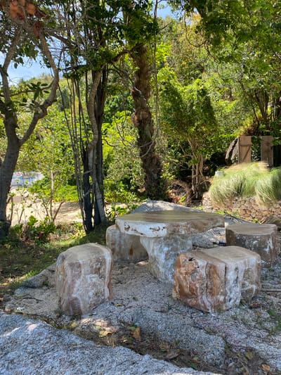 The image depicts a natural outdoor setting with a rock bench composed of large, stacked marble-like stones. The bench sits on a rocky terrain mixed with small pebbles and sparse vegetation. Behind the bench, a dense thicket of trees and shrubs forms a green backdrop. A path leads away from the bench, disappearing into the vegetation. The location appears to be near Ban Haad Rin, Thailand, inferred from the typical vegetation. No people are visible in the shot. The focus is on the natural environment and the constructed rock bench within it.