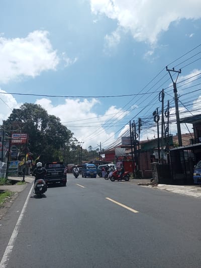 The image depicts a street scene in what is likely a residential or commercial area. The sky is mostly blue with scattered white clouds. A street with a yellow dividing line is visible, with a motorcycle and a dark-colored SUV traveling in the same direction. The motorcycle rider is wearing a helmet and a jacket with the word "Grab" on it. Other vehicles, including a blue van, are present in the distance. Buildings line the right side of the street, with signs that suggest the presence of shops or businesses. One prominent sign is for "PRONES APOTEK." The left side of the street has signage including an "Alfamart" logo. Power lines are strung across the scene. The general environment suggests a warm and sunny day. The specific location within Kioumaknao 2, Laos is unidentifiable without more information.