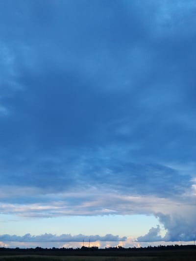 The image shows a wide-angle view of a cloudy sky over a rural landscape. The sky is predominantly a deep blue, with variations in cloud density and color. Near the horizon, the clouds are lighter, with hints of white and pink, suggesting a sunset or sunrise. The lower portion of the image depicts a dark silhouette of trees and possibly some structures against the brighter sky. A faint line of what appears to be utility poles can be seen along the horizon on the right side. The foreground consists of a dark band that could be fields or vegetation. The overall impression is of an open, natural environment with dynamic cloud formations. There are no people or animals visible in the image.
