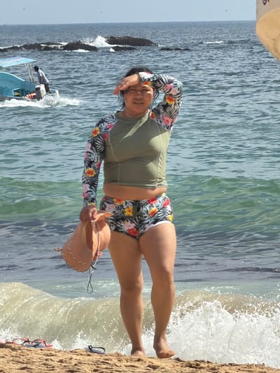 A woman in a green rash guard with floral sleeves and matching floral swim bottoms stands on a sandy beach. She is holding a straw hat in one hand and has the other hand on her forehead. The ocean is behind her, with waves crashing onto the shore. A small boat is visible in the distance to the left. Red flip-flops are next to the woman's feet.