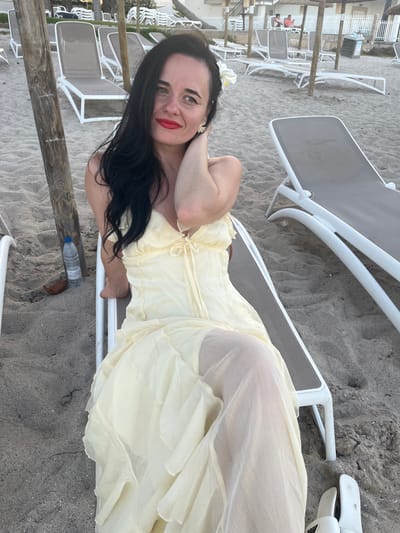A woman with dark hair and red lipstick sits on a lounge chair on a sandy beach. She is wearing a light yellow dress and a white flower in her hair. In the background, there are more lounge chairs, umbrellas, and buildings. A bottle of water and a piece of coconut are on the sand near her. The scene appears to be in Platja D'alcúdia, Spain, indicated by the presence of beach amenities consistent with that location. The lighting suggests it is either early morning or late afternoon.
