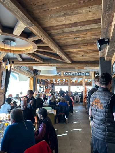 The image depicts a busy, warm interior scene, likely a restaurant or bar in a ski resort. Numerous people are gathered, mostly seated at tables, engaged in dining, conversation, or waiting. The setting is characterized by extensive use of natural wood, forming the walls, ceilings, and prominent structural beams, creating a rustic, chalet-like atmosphere.
Bright natural light streams through large windows in the background, illuminating the space and suggesting it is daytime. This light casts distinct reflections on the floor. Various lighting fixtures are visible, including recessed spotlights, modern pendant lights with exposed bulbs, and large circular ceiling lights with diffusers. Some sections of the walls feature decorative black and white patterned tiles.
A notable detail is the orange text "L'ETINCELLE VAL D'ISÈRE" clearly visible on the back of a grey puffer vest worn by a person standing in the mid-ground. This text strongly indicates the location is Val-d'Isère, France, and "L'Etincelle" is likely the name of the establishment. The attire of the people and the wooden decor further support the context of a ski resort setting. The overall mood appears lively and social.