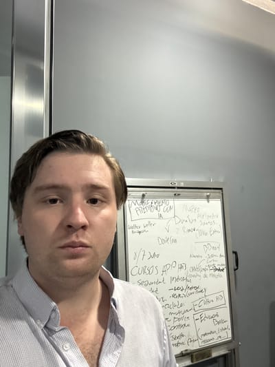 A young Caucasian man with light brown hair and a fair complexion is taking a selfie in an indoor setting. He is wearing a light blue and white checkered button-down shirt with the top button undone. His expression is neutral, and he is looking directly at the camera.

Behind him, a whiteboard is visible, covered in handwritten notes in Spanish. The writing appears to be a mix of project tasks, names, and dates. Some legible phrases include "MANTENIMIENTO PREDICTIVO CON IA" (Predictive Maintenance with AI), "Walter Weller" (likely a name), "Doble Coin" (likely a cryptocurrency name), "CURSOS APP HTS" (App Courses HTS), and "Educación Básica" (Basic Education). There are also names like "Alejandro Santos" and "Juan Extra".

The environment appears to be an office or workspace, with a metallic door or partition to the left of the man. The lighting suggests an indoor setting, possibly artificial light given the reflection on the metallic surface. The background wall is a neutral gray. The presence of a whiteboard with detailed notes implies a work or study-related context. The image does not contain any direct indicators to pinpoint the exact location within Cuautitlán Izcalli, Mexico, beyond the inferred context of a workplace or educational facility.
