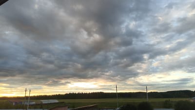 The image depicts a rural landscape under a cloudy sky, likely taken in Ankudínovo, Russia. The sky is filled with dense, dark gray clouds interspersed with lighter patches of white and hints of yellow and pink, suggesting either an impending storm or a sunset/sunrise. In the distance, a dark line of trees forms a forest against the horizon. In the foreground, there are some man-made structures. To the left, there are the roofs of buildings, including what appears to be a greenhouse. Several utility poles with wires are visible in the middle ground. The overall scene is one of a natural environment with some signs of human habitation.