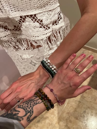 Two human arms are positioned in the center of the frame. The arm on the left is adorned with a stack of beaded bracelets in shades of white, green, and black. The hand has a gold ring on the middle finger. The arm on the right features a tattoo of a quadrupedal animal, as well as two beaded bracelets in tones of pink and brown-green. A ring is visible on the pinky finger of this hand. A white, intricately crocheted garment is visible to the left of the arms. The background is out of focus but appears to be a tiled floor and part of a wall. The image appears to have been taken indoors. The location in the city of Puçol, Spain is undetermined.