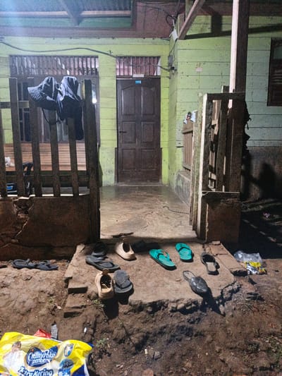 The image depicts the entrance to a simple, weathered wooden building in Meunasahuket, Indonesia. The scene is set at dusk or night, indicated by the artificial lighting casting shadows and the muted colors. A wooden porch with a railing extends to the left, with two dark-colored jackets or clothing items hung over it.
In front of the building's entrance, which features a dark wooden door, several pairs of footwear are scattered on a concrete pathway and the surrounding dirt. These include flip-flops in teal and dark colors, casual sandals, and a pair of light-colored slip-on shoes. The ground is uneven and appears to be a mix of dirt and worn concrete.
To the right of the entrance, another section of wooden fencing stands, and a dark shadow is cast on the light green wooden wall of the building. In the foreground, a bright yellow plastic bag is partially visible, with some text printed on it, including "CreamyPoko pants" and "Extra Kering". The overall impression is one of a humble, lived-in dwelling, likely in a rural or semi-rural area, with everyday items left outside. There are no people visible in the image.
