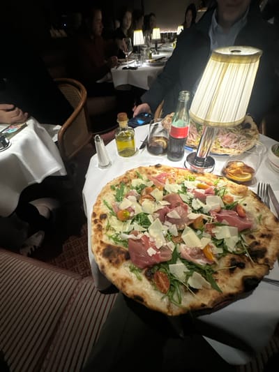 A restaurant table features two pizzas, one with prosciutto, arugula, parmesan, and tomatoes. Other items include a Coca-Cola bottle, an olive oil bottle, a salt shaker, and lemon slices. People are seated at the table with table lamps providing light. A woven chair is near the table, and a person's legs and shoes are visible. The tablecloth is white and there's a red striped pattern on the chair next to the table.