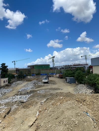 This is an outdoor, high-angle, eye-level shot of a construction site in Baraki, Algeria, on a clear day with scattered white clouds. The foreground is dominated by piles of dirt and debris, suggesting demolition or excavation. A white excavator is positioned in the middle ground, facing to the right. Behind it, there are piles of broken concrete and rubble. Further back, a large building complex is under construction, covered in green netting and scaffolding. Several tall construction cranes are visible, with two prominent green cranes in the distance. The environment appears to be an urban setting, with evidence of ongoing development.