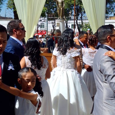 Cerro Colorado, Peru. An outdoor wedding ceremony takes place under a white tent. The bride and groom stand in the background. In the foreground, a man in a blue suit stands next to a young girl in a white dress. They are holding hands. In the background, a tall building is visible behind some trees.