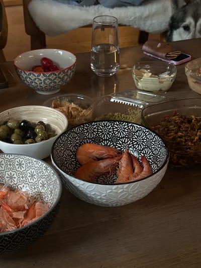 The image shows a wooden table laden with various food items and containers, likely in a domestic setting. There are bowls containing shrimp, olives, and cherry tomatoes. Additionally, there are clear containers holding spreads, possibly pate or dip, and a jar of fried onions. A glass of water and a purse are also present. A portion of a white dog is visible in the left side of the frame. Location in the city of La Couarde-sur-Mer, France is not evident from the image.