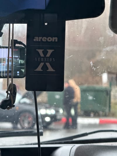 The image provides a view from inside a vehicle, looking out through a windshield that appears wet and somewhat dirty. The original image is oriented vertically, showing elements both inside and outside the vehicle.
In the foreground, hanging inside the car, is a black air freshener with the brand "areon" and "VERSION X" clearly visible in white text. Below it, a smartphone is mounted, likely functioning as a dashcam or recording device, with its screen displaying the time "02:23". A small, dark, perhaps decorative, object hangs by a cord to the right of the phone. Part of the vehicle's interior, possibly a sun visor, is visible at the top left.
The outdoor scene, viewed through the rain-streaked glass, appears blurry and dimly lit, suggesting overcast conditions or low light, consistent with the early morning time shown on the phone (02:23). In the mid-ground, a red and white striped barrier or pole is visible on the right, indicating a possible checkpoint or restricted area. Green dumpsters or containers are visible further in the background.
A dark-colored car with distinctive circular headlights is seen from the rear in the center-right of the frame. Behind this vehicle, a person wearing a light-colored top is bent over, their activity unclear due to blur and obstruction. Another person, dressed in darker clothing, is partially visible nearby. The scene suggests the vehicle from which the photo is taken is stopped or waiting.
The visible text includes "areon", "VERSION X", and the digital time "02:23". The location context for this scene is Klenovskoe, Russia.