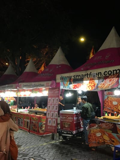 The image shows a night market in Kecamatan Batu, Indonesia. The market is located under a series of white and pink tents with the words "Smartfren" on them, which is a mobile network provider. It is located at a busy street with a concrete sidewalk. There are various food stalls with different menu items on display. There are several people present, including a few customers browsing the different options, and a few people working at the stalls. There are also numerous food items displayed and on sale. The tents are lit up by overhead lights, and there is a single moon in the sky. There are multiple tables and chairs set up throughout the scene. There are several food items on the display, including: fried chicken, french fries, onion rings, fried shrimp, and various beverages. There is a single ice cream cart with a sign that reads "Ngopi Candu". The people are dressed casually in everyday attire. The market appears to be bustling with activity, with several people walking around and looking at the different stalls. There is a single trash can in the lower right of the frame.